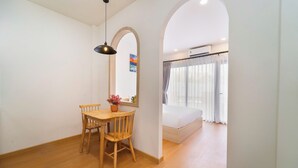 A room with a wooden dining table, two chairs, a hanging light fixture, a bed with a white mattress, and a window with curtains. - SORA HOTEL & APARTMENT (Ho Chi Minh City)