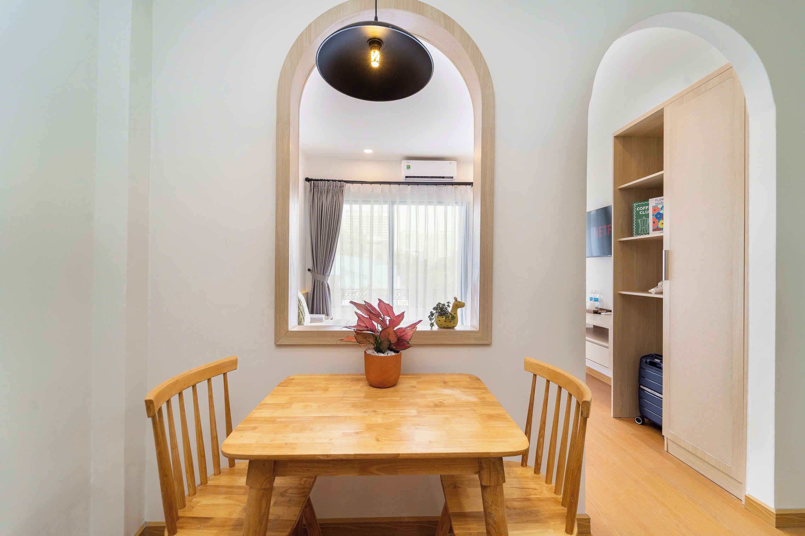 A dining area with a wooden table and chairs, a potted plant, a pendant light, a window with curtains, and a bookshelf.