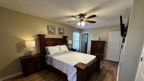 A bedroom with a wooden bed, a ceiling fan, a nightstand with a lamp, a dresser, and a door. - Oak Breeze, Coastal get away between 2 Paradises (Gulf Breeze)