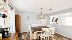 A dining area with a wooden table and white chairs, a chandelier, and a television on the wall. - Oak Breeze, Coastal get away between 2 Paradises (Gulf Breeze)