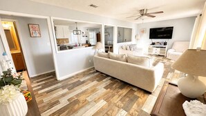 A modern living room with a sofa, a TV, and a dining area with a kitchen in the background. - Oak Breeze, Coastal get away between 2 Paradises (Gulf Breeze)
