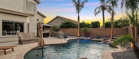 A swimming pool with a curved edge, surrounded by a patio area with outdoor furniture and palm trees.