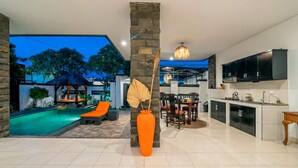 A modern kitchen with black cabinets and a large TV, a dining area with a chandelier, and a pool area with lounge chairs. - Sunbeji Villa (Seminyak)