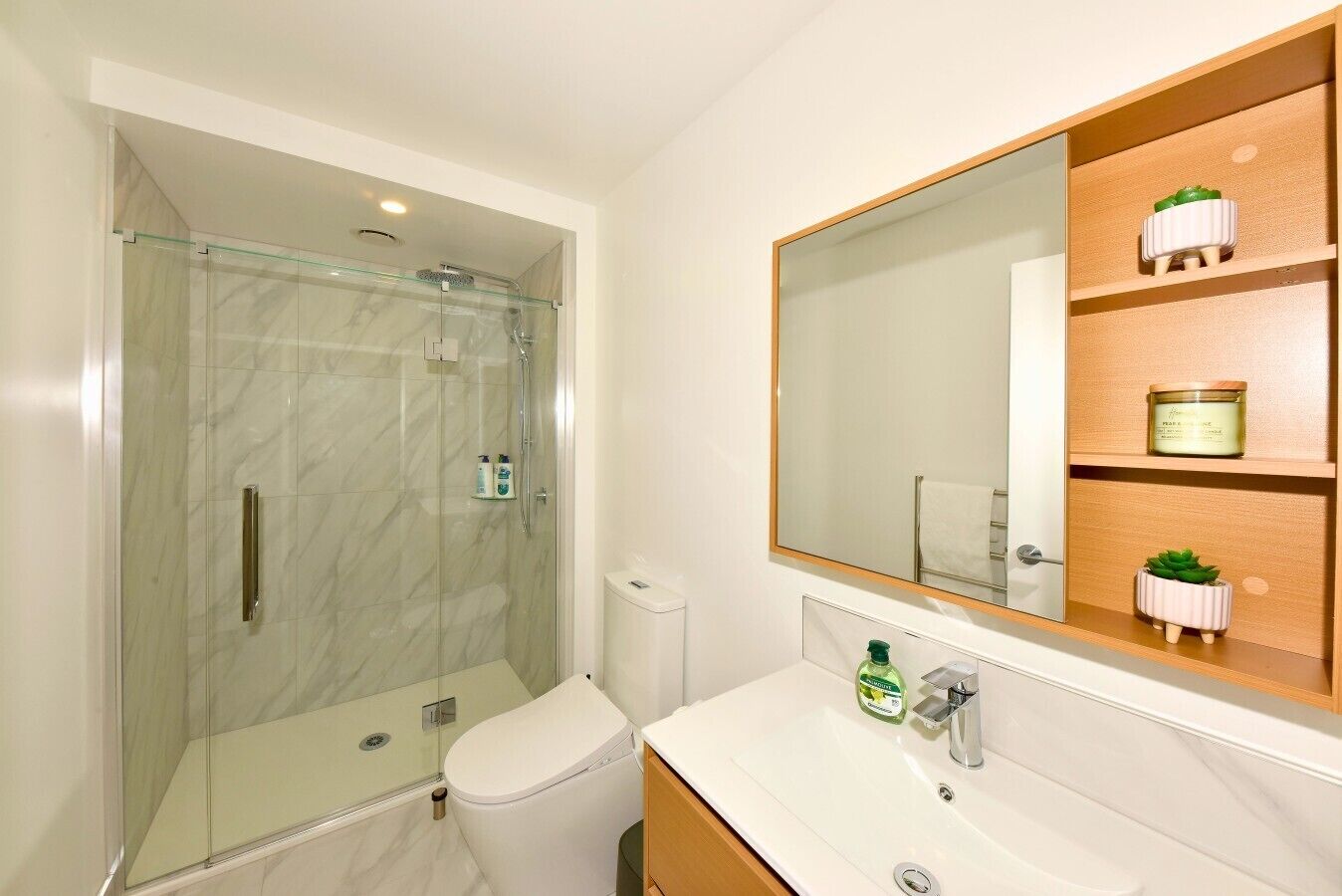 Une salle de bain moderne avec une douche à parois de verre, des toilettes blanches, un lavabo avec un miroir et une étagère contenant des articles de toilette.