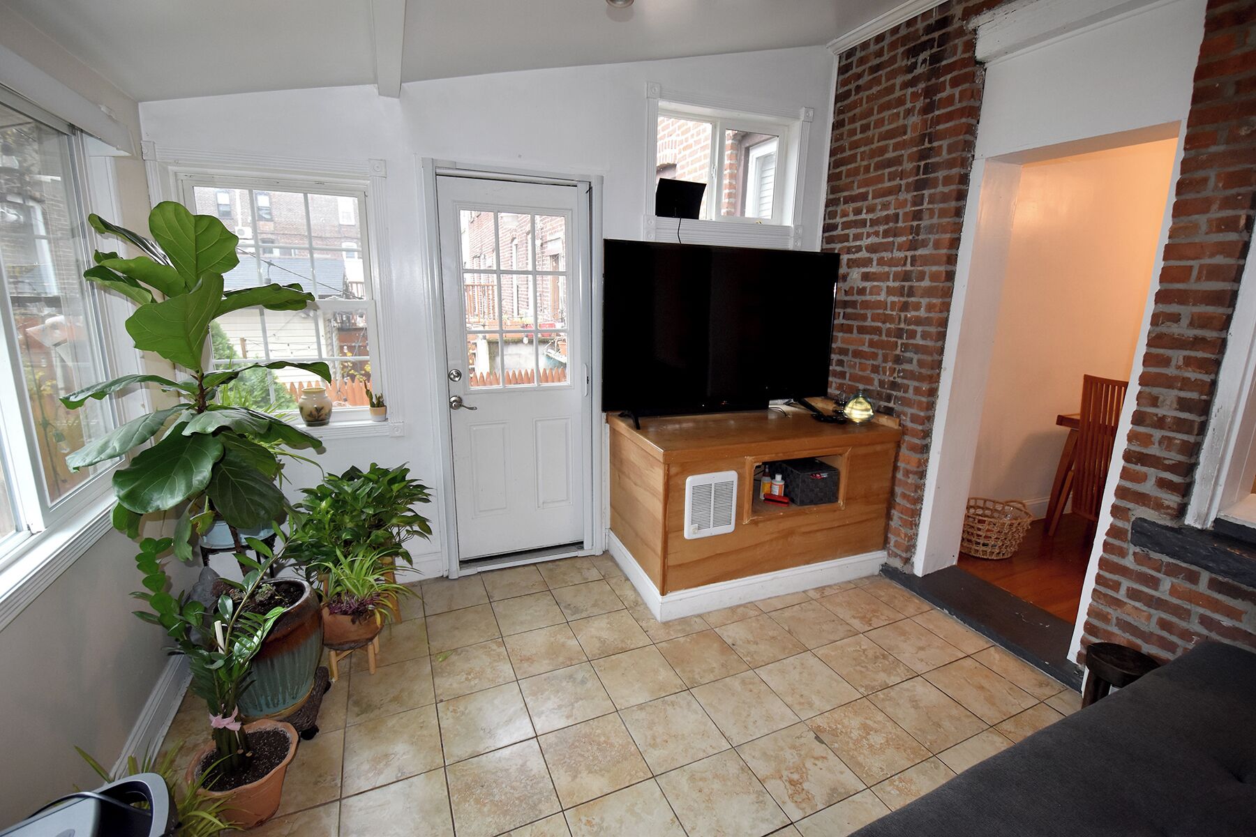 Una sala de estar con televisor, una puerta, pared de ladrillo y plantas en macetas.
