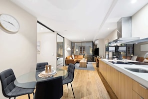 Eine moderne Küche mit einer zentralen Kochinsel, einem Essbereich mit Glastisch sowie ein Wohnbereich mit Sofa und Couchtisch.
