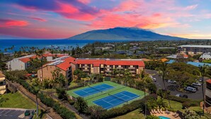 Een hotel met een tennisbaan, omringd door groen en een berg op de achtergrond.