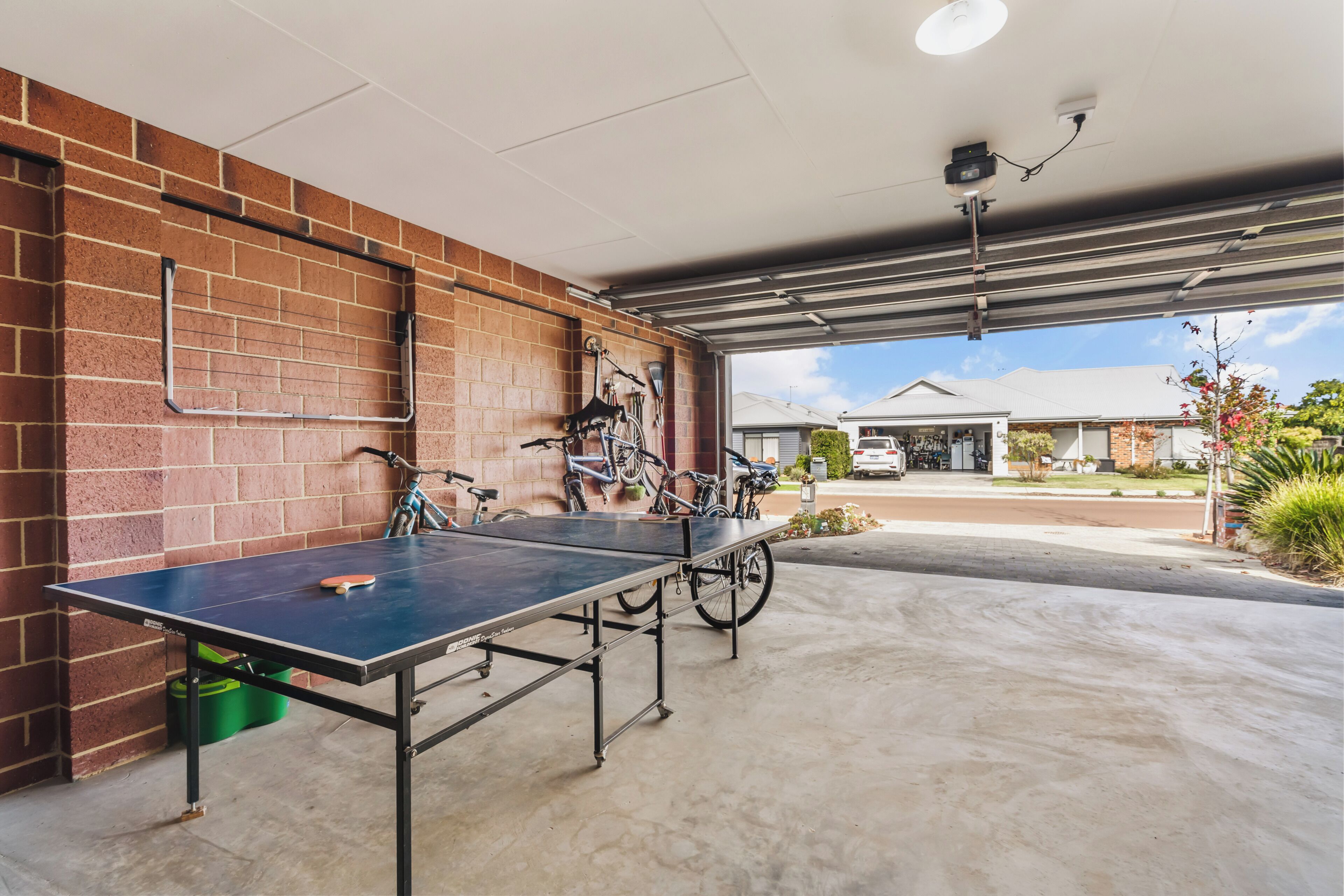 Une table de ping-pong dans un garage ouvert, avec des vélos et une maison en arrière-plan.