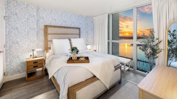 Ein Schlafzimmer mit einem großen Bett, einem Nachttisch, einem Beistelltisch, einem Sessel, Blick auf den Sonnenuntergang und einem Balkon.