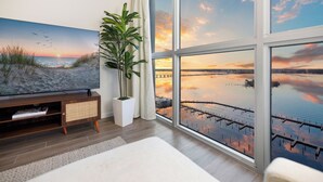 Ein Zimmer mit großem Fenster, einem Sonnenuntergangsbild, einer Pflanze und einem Fernseher auf einem Holzständer.