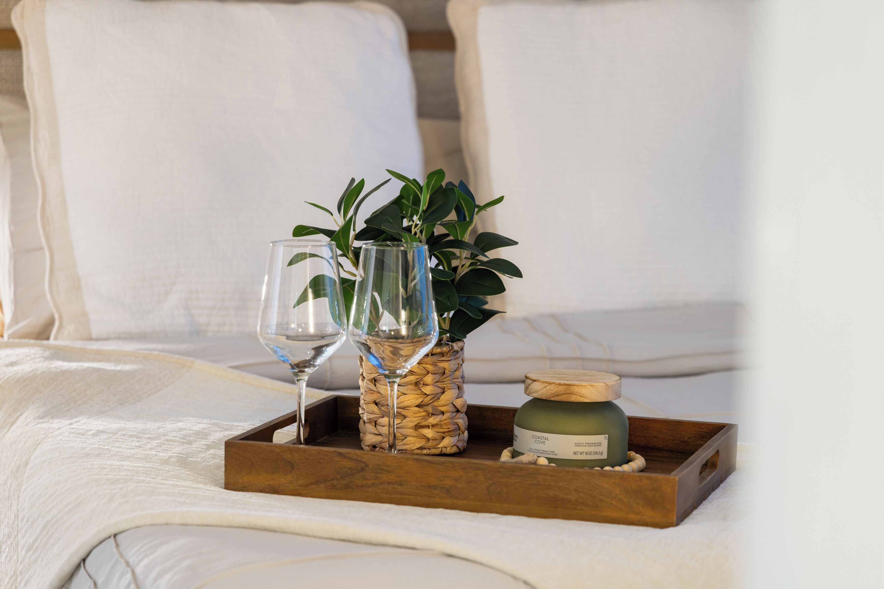 A wooden tray with a glass, a woven container with green plants, and a candle on a bed.
