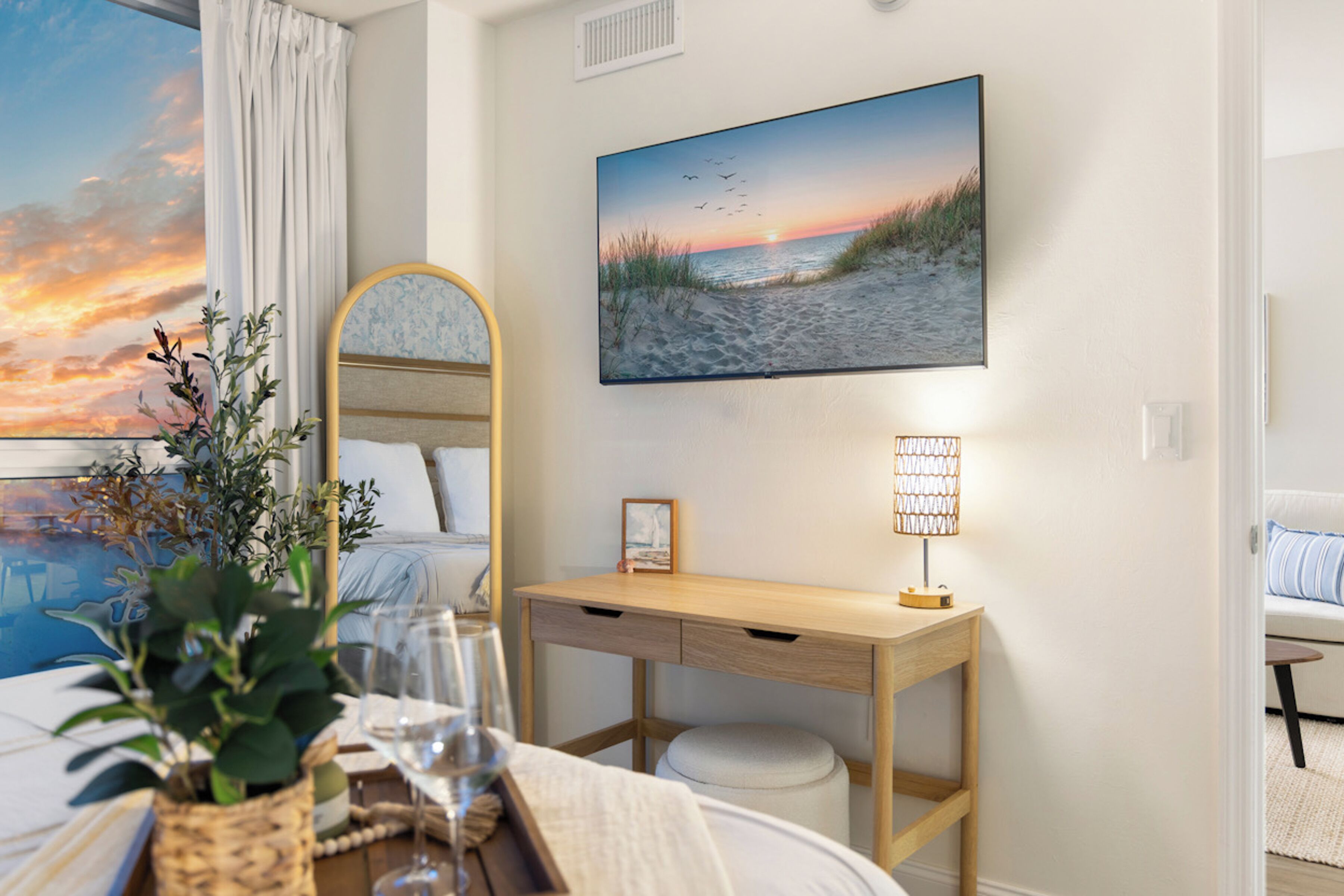 Una habitación de hotel con cama, escritorio, lámpara, un cuadro de una playa y ventana con cortinas.