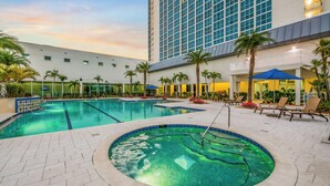 Ein Hotelpoolbereich mit einem großen Gebäude im Hintergrund, Palmen und Liegestühlen.