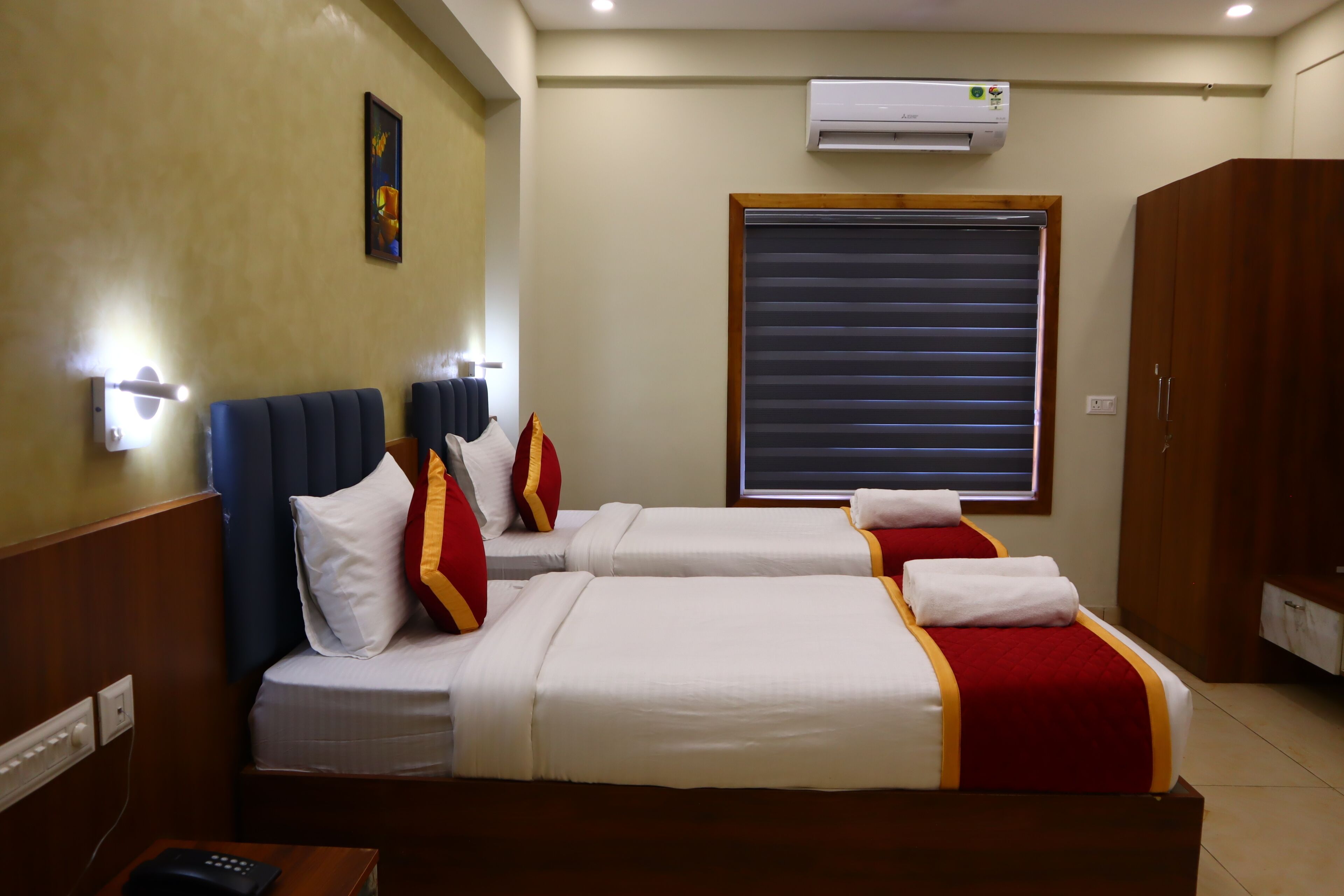A hotel room with two beds, a window with blinds, an air conditioner, and a wall-mounted light fixture.