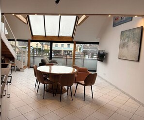 Un espace repas moderne avec une table ronde, plusieurs chaises, un canapé et une grande fenêtre donnant sur des immeubles.
