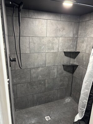 Eine Dusche mit grauen Fliesen, einem Duschhandstück und einem schwarzen Regal.