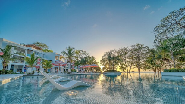 Una piscina con sillones de descanso y sombrillas, rodeada de palmeras y un edificio con balcones.