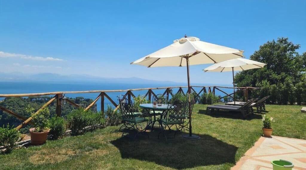 Ein Sitzbereich im Freien mit Tisch und Stühlen, einem großen Sonnenschirm und Blick aufs Meer.
