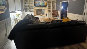 Una sala de estar con un sofá seccional negro, una chimenea, un televisor montado encima y una estantería llena de objetos variados.
