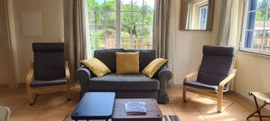 Ein Wohnzimmer mit grauem Sofa, zwei Sesseln, einem Couchtisch und einem Fenster mit Blick ins Grüne.
