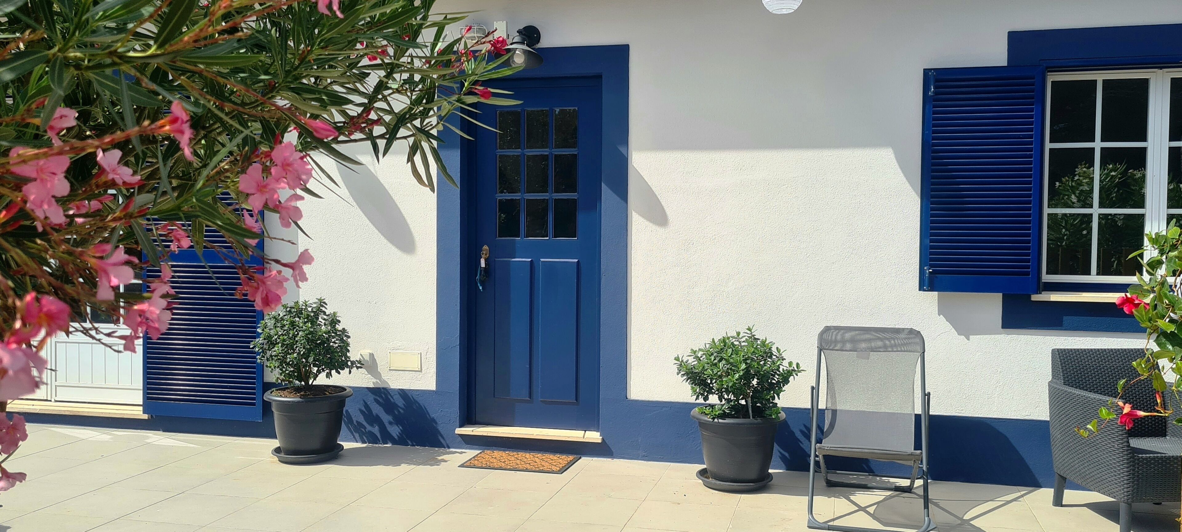 Un bâtiment avec une porte bleue, deux plantes en pot, une chaise et une fenêtre avec des volets bleus.