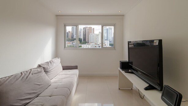 A modern living room with a sofa, a flat-screen TV, and a city view through large windows. - Ap Bela Vista c Portaria 24h Paulista e Masp (São Paulo)
