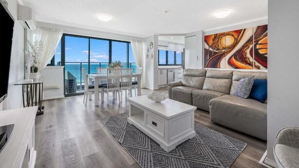 A modern living room with a large window, a sofa, a coffee table, and a dining area. - Sunny Coast Escapes Maroochydore (Maroochydore)