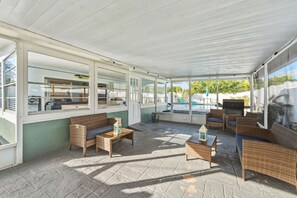 Ein überdachter Außenbereich mit Korbmöbeln, Steinboden und großen Fenstern mit Blick auf einen Pool.