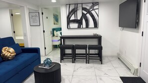 A modern living room with a blue sofa, a black coffee table, a black bar with stools, a large abstract painting, and a TV on the wall. - Adorable 2-br apt Pet-Friendly .Fast WiFi .Near Airport & LIRR (Central Islip)