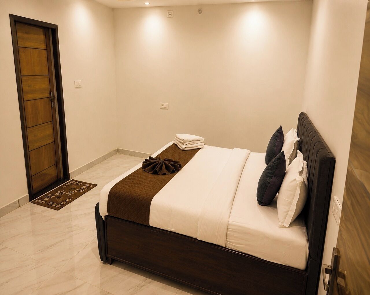 Una habitación de hotel con cama, puerta de madera, alfombra pequeña y toma de corriente.