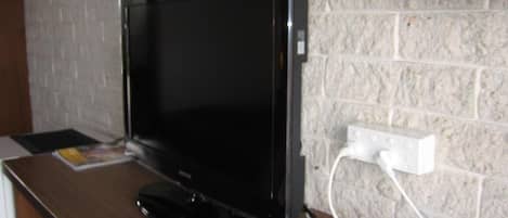 Televisor sobre superficie de madera, con control remoto y un decodificador, conectado a un enchufe blanco en una pared con textura.