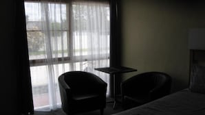 Et svagt oplyst hotelværelse med en seng, to stole, et lille bord og et vindue med lette gardiner.