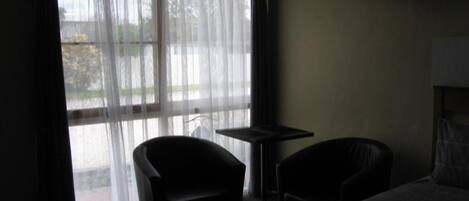 A dimly lit hotel room with a bed, two chairs, a small table, and a window with sheer curtains.