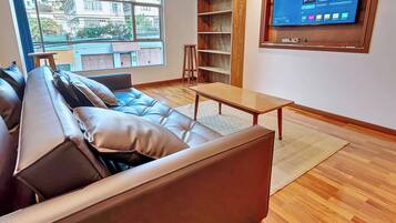 Una sala de estar moderna con un sofá de cuero, una mesa de centro de madera, un televisor montado en la pared y vistas al exterior a través de un amplio ventanal.