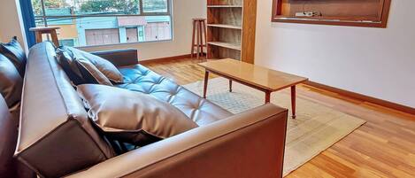 Un salon moderne avec un canapé en cuir, une table basse en bois, un téléviseur fixé au mur et une vue sur l’extérieur grâce à une grande fenêtre.