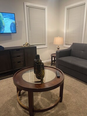 Ein Wohnzimmer mit rundem Glastisch, einer Couch, einem an der Wand montierten Fernseher und zwei Fenstern mit Jalousien.