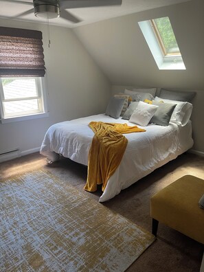Ein Schlafzimmer mit Bett, Kissen, Decke, einem Fenster, einem Oberlicht und einem Deckenventilator.