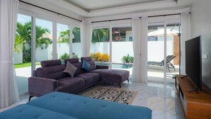 A modern living room with a sectional sofa, a flat-screen TV, and large sliding glass doors leading to a pool area. - Orchid Paradise Homes OPV429 (Hin Lek Fai)