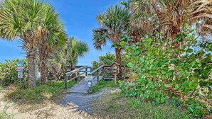 A sandy pathway leading to a wooden bridge with signs, surrounded by palm trees and greenery. - Cozy 1BR Ground Floor Apartment Steps to Beach (Cocoa Beach)