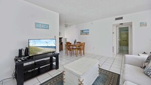 A living room with a TV, a dining table, and chairs. There is a sign on the wall that says 'BEACH'. - Cozy 1BR Ground Floor Apartment Steps to Beach (Cocoa Beach)