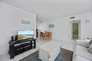 Ein Wohnzimmer mit Fernseher, Esstisch und Stühlen. An der Wand hängt ein Schild mit der Aufschrift 'BEACH'.