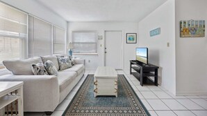 A living room with a sofa, a coffee table, a TV, and a door. - Cozy 1BR Ground Floor Apartment Steps to Beach (Cocoa Beach)