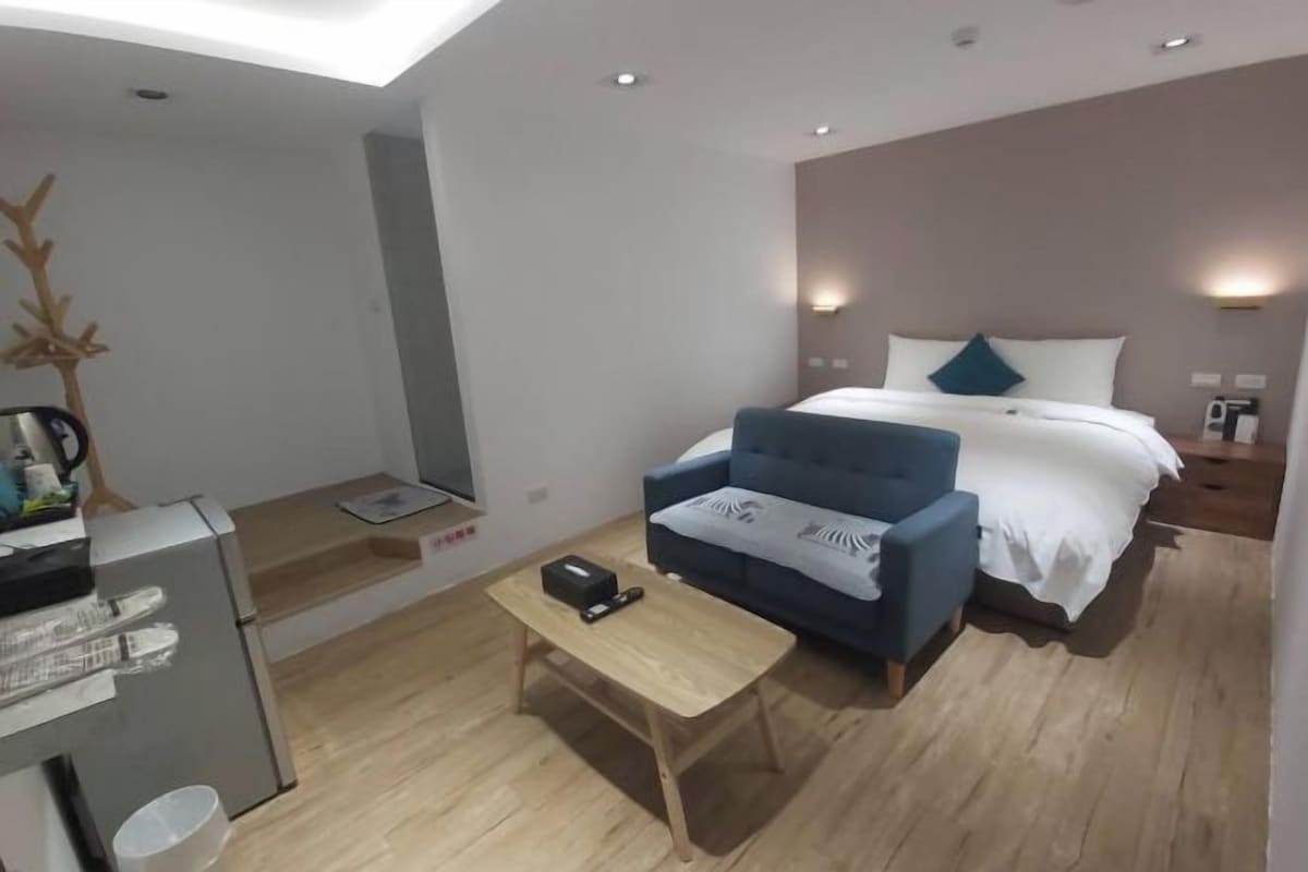Una habitación de hotel moderna con una cama, un sillón azul, una mesita de madera y un escritorio pequeño con una computadora y una taza.
