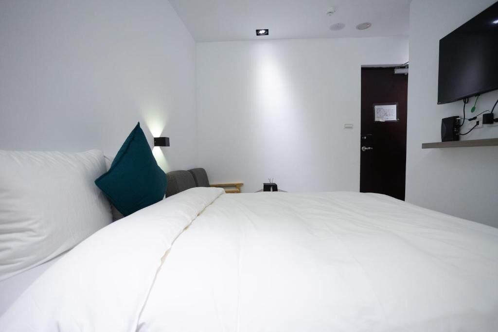 Ein Hotelzimmer mit einem großen Bett, einem flachbildfernseher an der Wand, einer Tür und einem kleinen Tisch mit einer Lampe.
