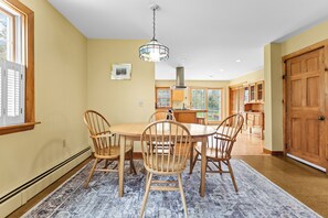A dining area with a wooden table and chairs, a hanging light fixture, and a view into the kitchen. - Acadia National Park Haven-New! (Mount Desert)