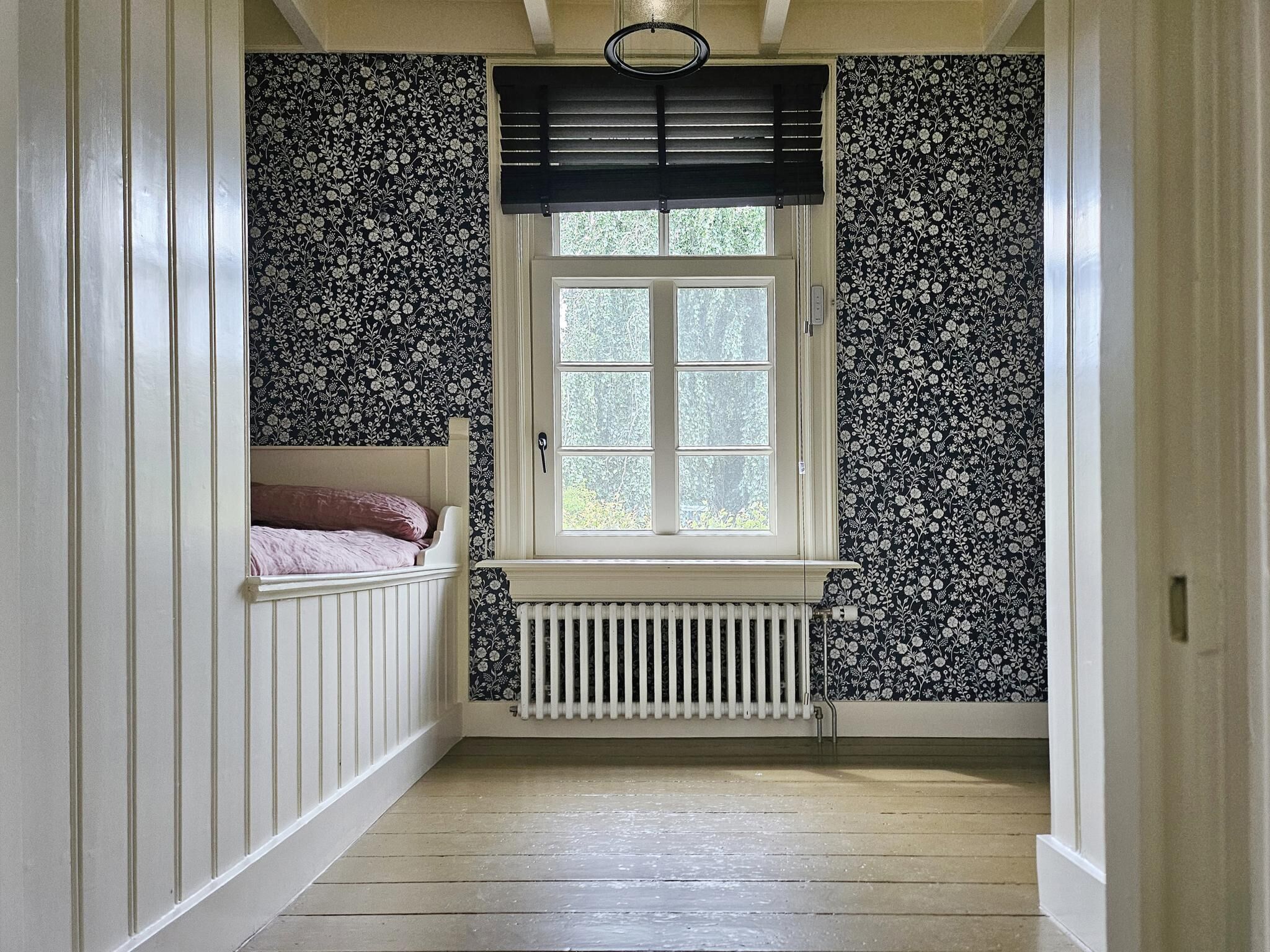 En smal gang med et vindue, en radiator og en seng med en lyserød pude.