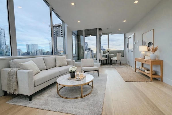 A modern living room with a sofa, coffee table, and chairs, large windows with a city view, and a dining area with a table and chairs. - Spectacular penthouse in a modernist, boutique concrete-and-glass building (Vancouver)