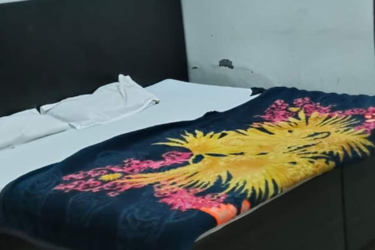 Una cama con colcha floral, almohada blanca y cabecera oscura en una habitación con paredes lisas y piso de baldosas.