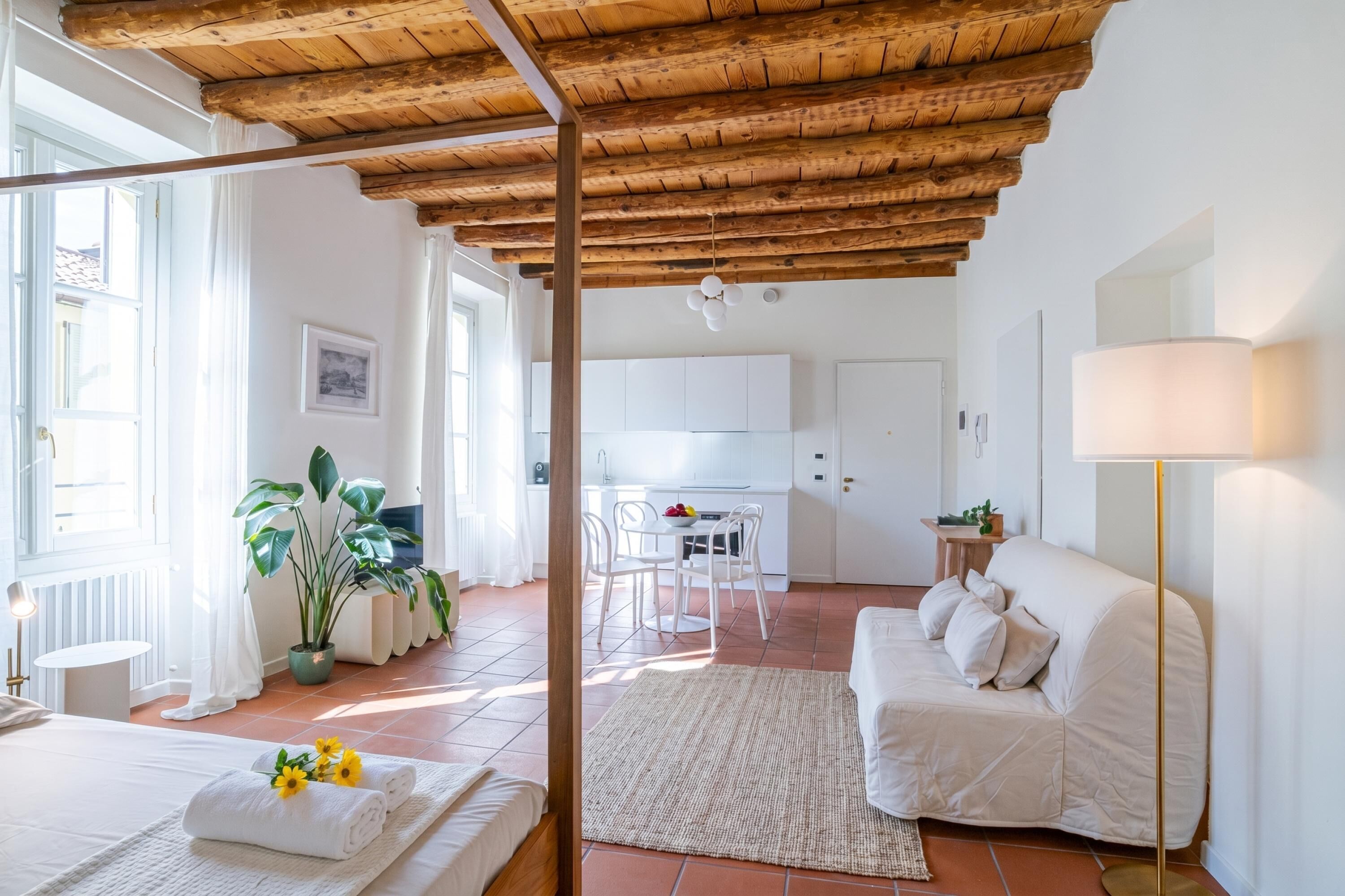 A room with a wooden ceiling, white walls, a bed with a white duvet, a white sofa, a dining table with chairs, and a kitchen area.