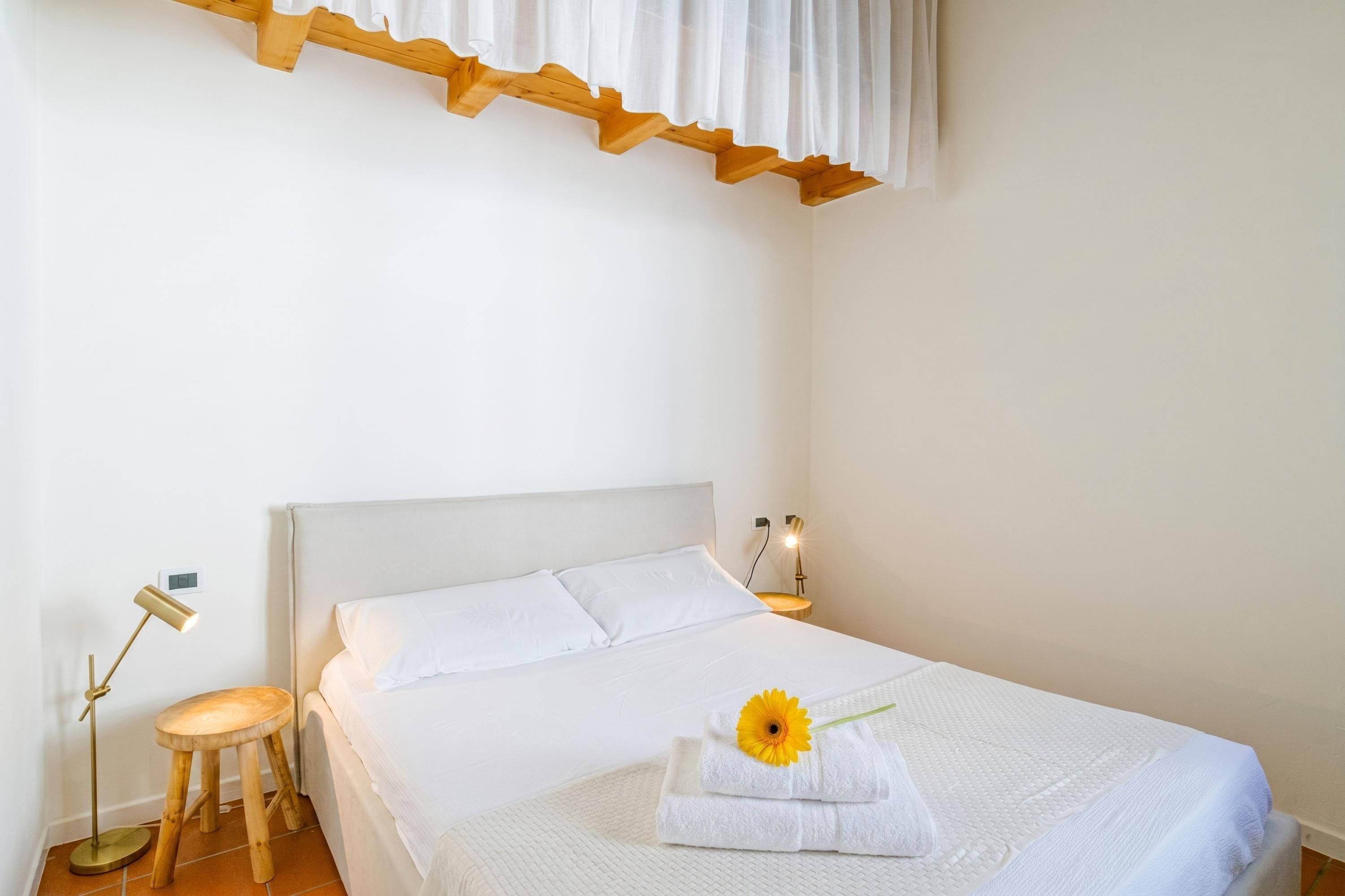 Un lit bien fait, avec du linge de lit blanc, un tabouret en bois, une lampe de chevet et une fleur posée sur le lit.